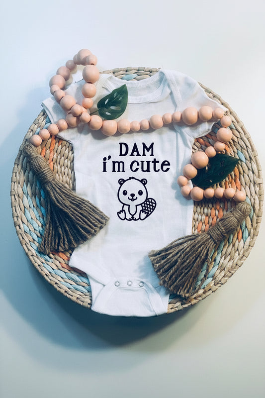 White baby onesie with 'DAM i'm cute' text and baby beaver graphic, surrounded by beads and leaves on a woven mat.
